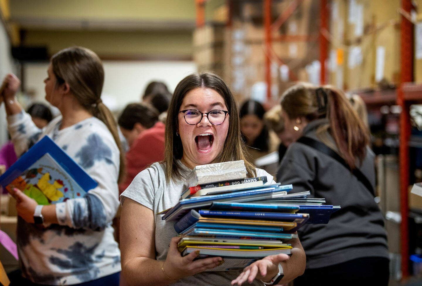A person with glasses joyfully holds a large stack of books in a warehouse setting, surrounded by others engaged in similar activities.